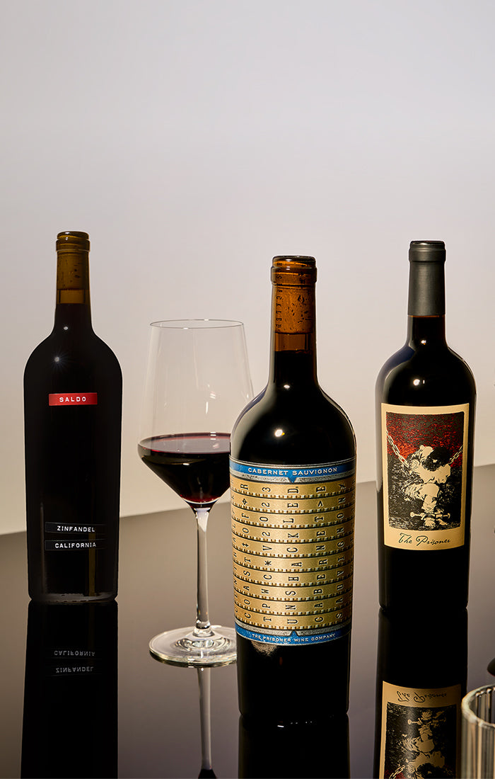 Three dark wine bottles and a filled wine glass arranged on a reflective surface against a plain light background. The center bottle has a gold-and-blue label, the left bottle has a red label, and the right bottle has a black-and-red label.