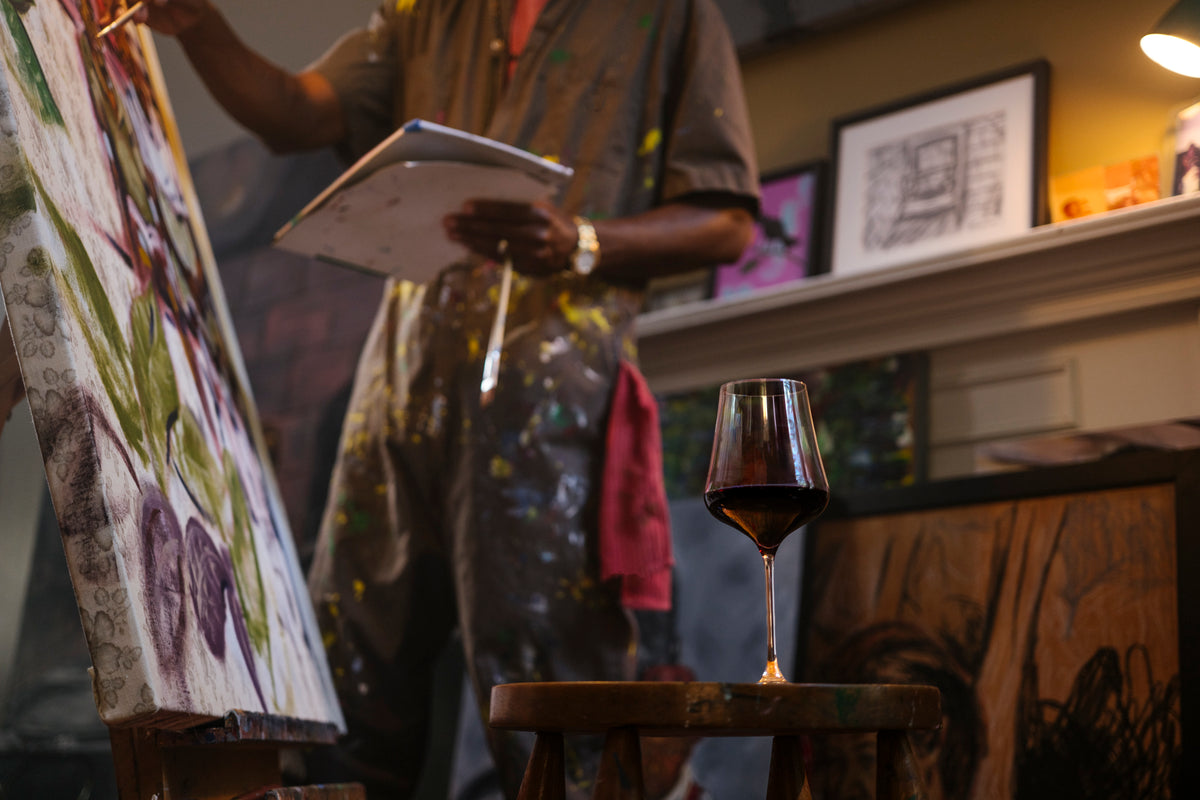 An artist in a paint-splattered apron stands at an easel holding a tablet, with a glass of red wine on a small wooden stool in the foreground and framed artwork in a warmly lit studio.