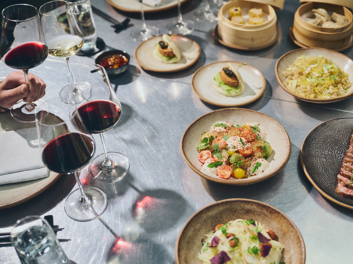 A high-angle view of a dining table filled with various dishes, including shrimp pasta, dim sum in bamboo steamers, and several wine glasses, with a hand holding a glass of red wine on the left.