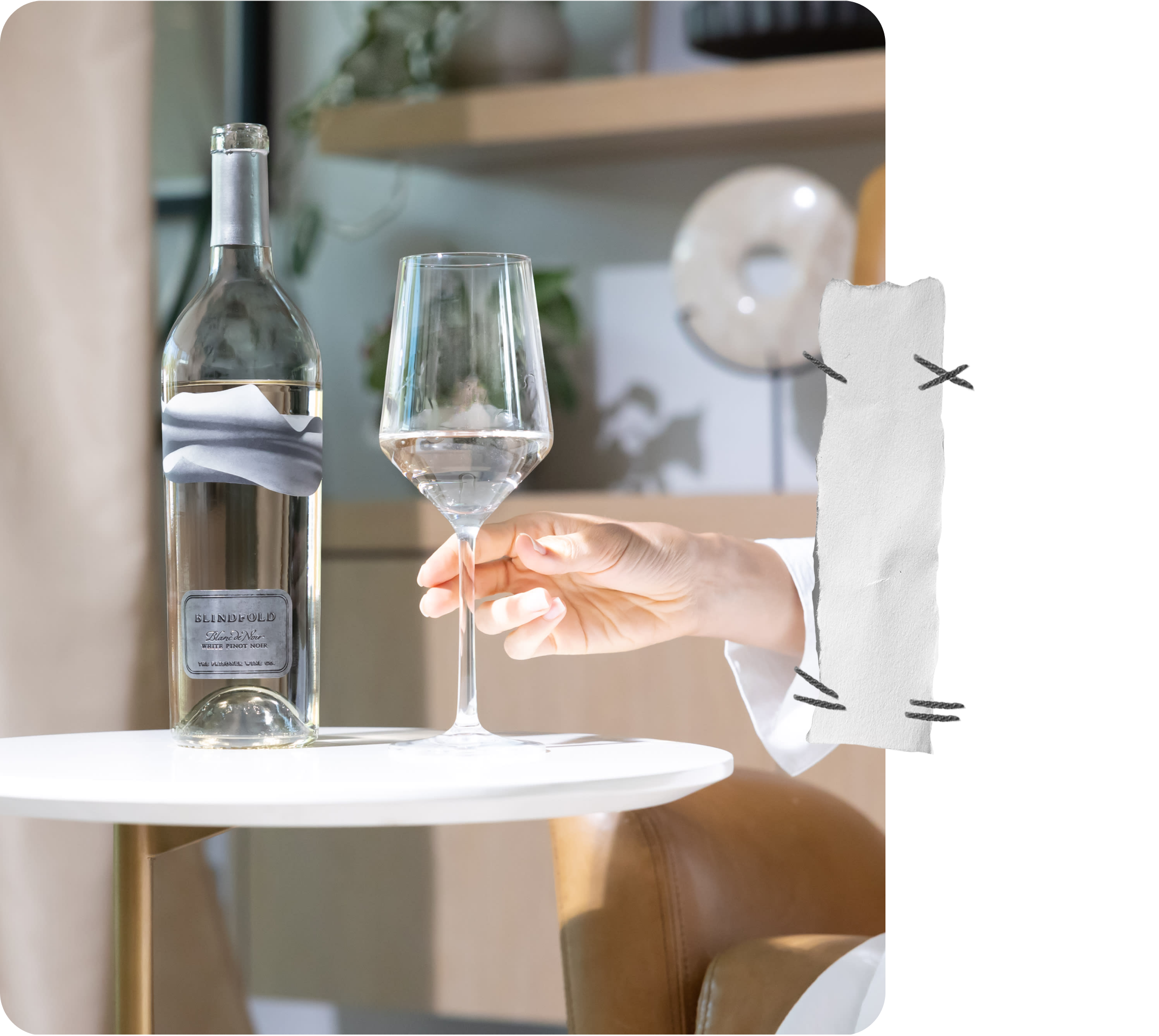 Person holding a wine glass next to a bottle on a small table with a blurred background
