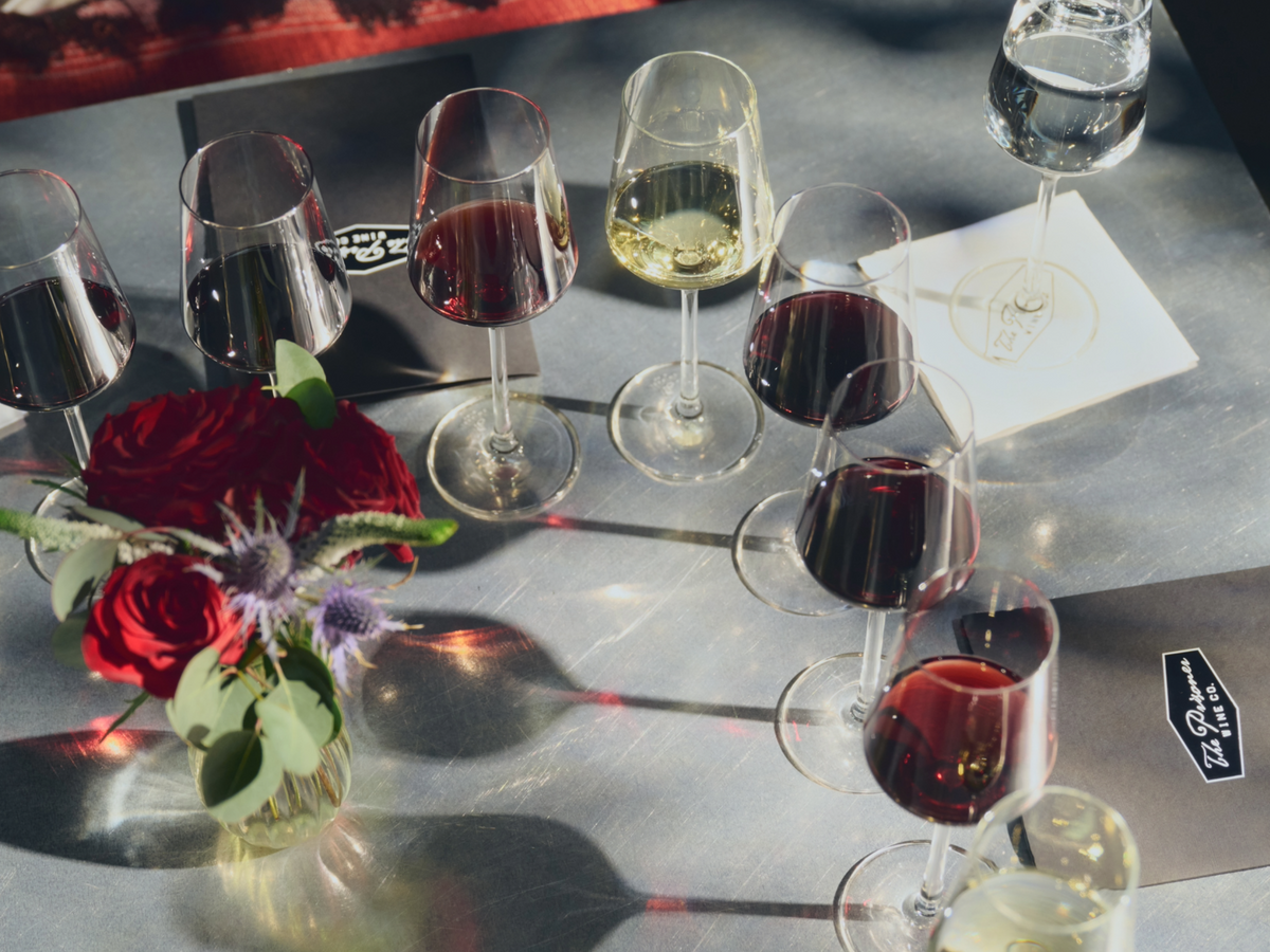 A tabletop arrangement of several wine glasses filled with red and white wine, alongside a small vase of red roses on a gray surface.