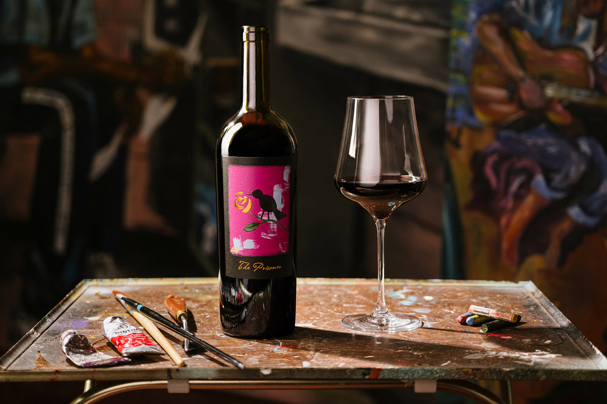 A bottle of red wine with a vibrant pink label featuring a black bird illustration stands next to a partially filled wine glass on a paint-splattered table, with paint tubes and brushes nearby against a blurred artistic background.