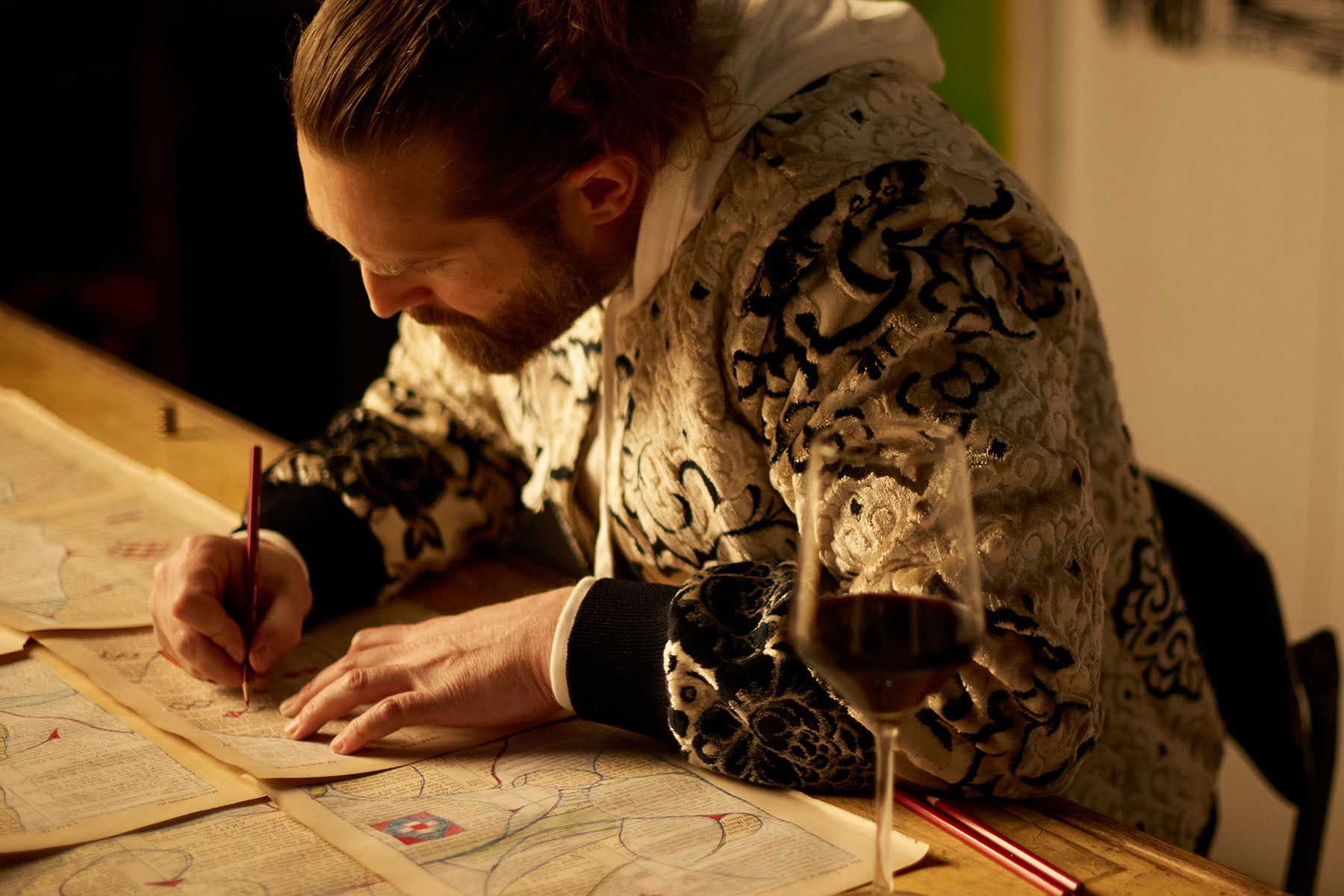 A man drawing on pages of writing next to a glass of red wine