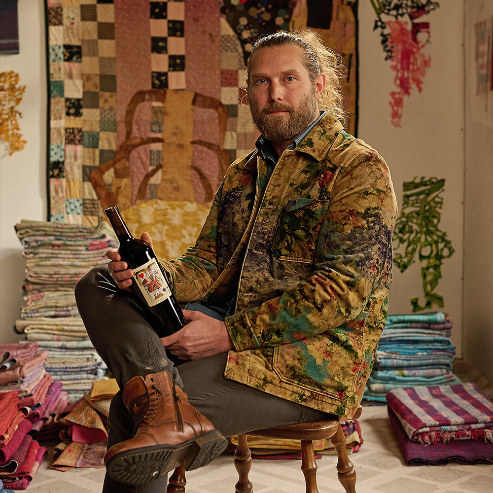 Man sitting on a chair holding a bottle of wine in a room with colorful fabrics and artwork.
