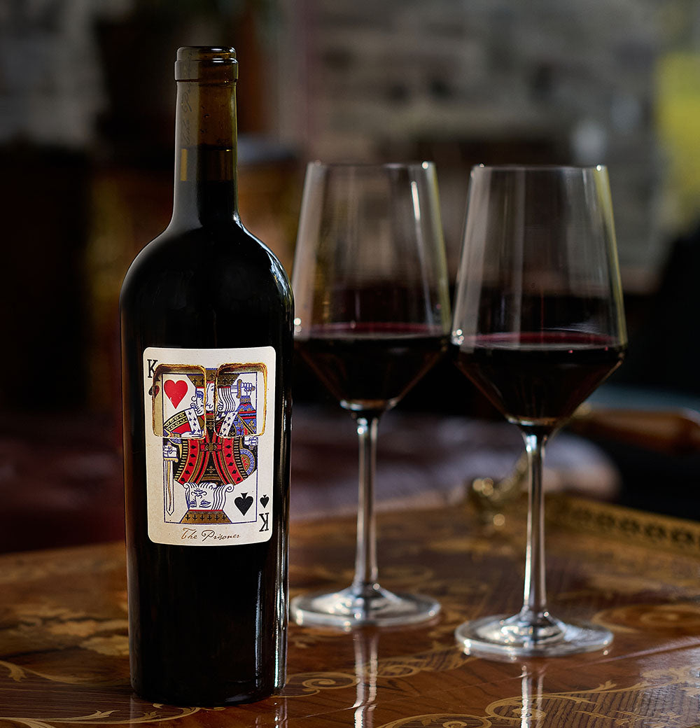 A bottle of The Prisoner x Corrections wine with a playing card label on a table with two glasses of red wine 