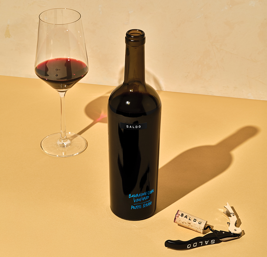 Bottle of Saldo Bavarian Lion Vineyard Petite Sirah on a yellow table with a wine glass and the cork on a corkscrew