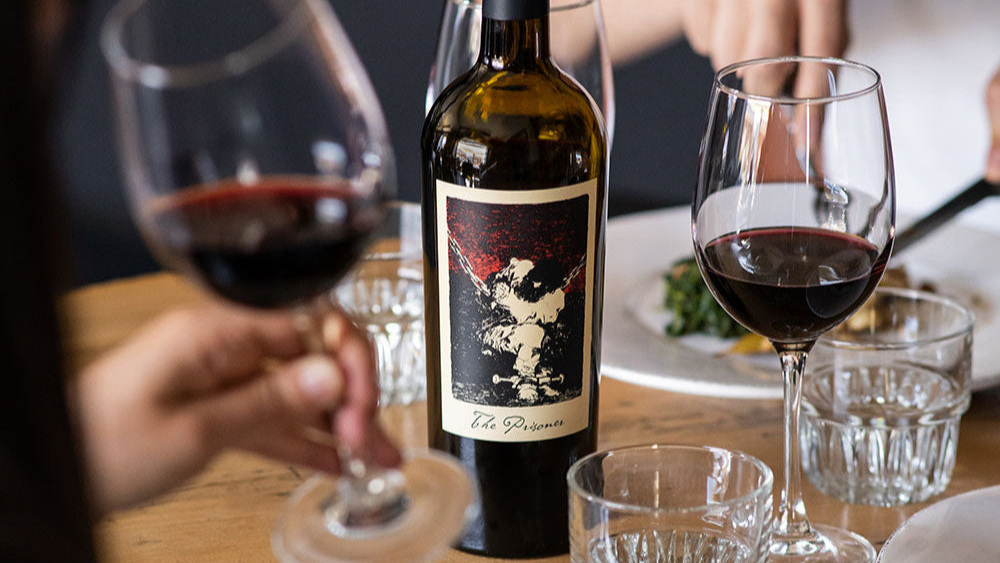 A bottle of The Prisoner Red Blend with two glasses of wine on a table surrounded by food 