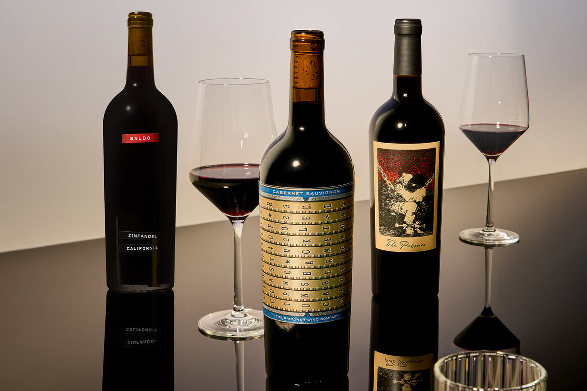 Three dark wine bottles with wine glasses on a reflective black surface against a light gray background. The center bottle has a large gold and blue label, the left bottle has a red label, and the right bottle has an illustrated label.