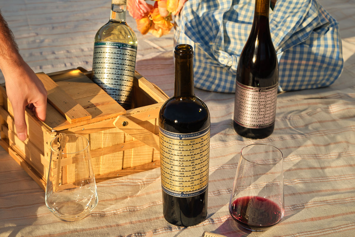 3 bottles of unshackled wines on a blanket with a basket and 2 glasses 