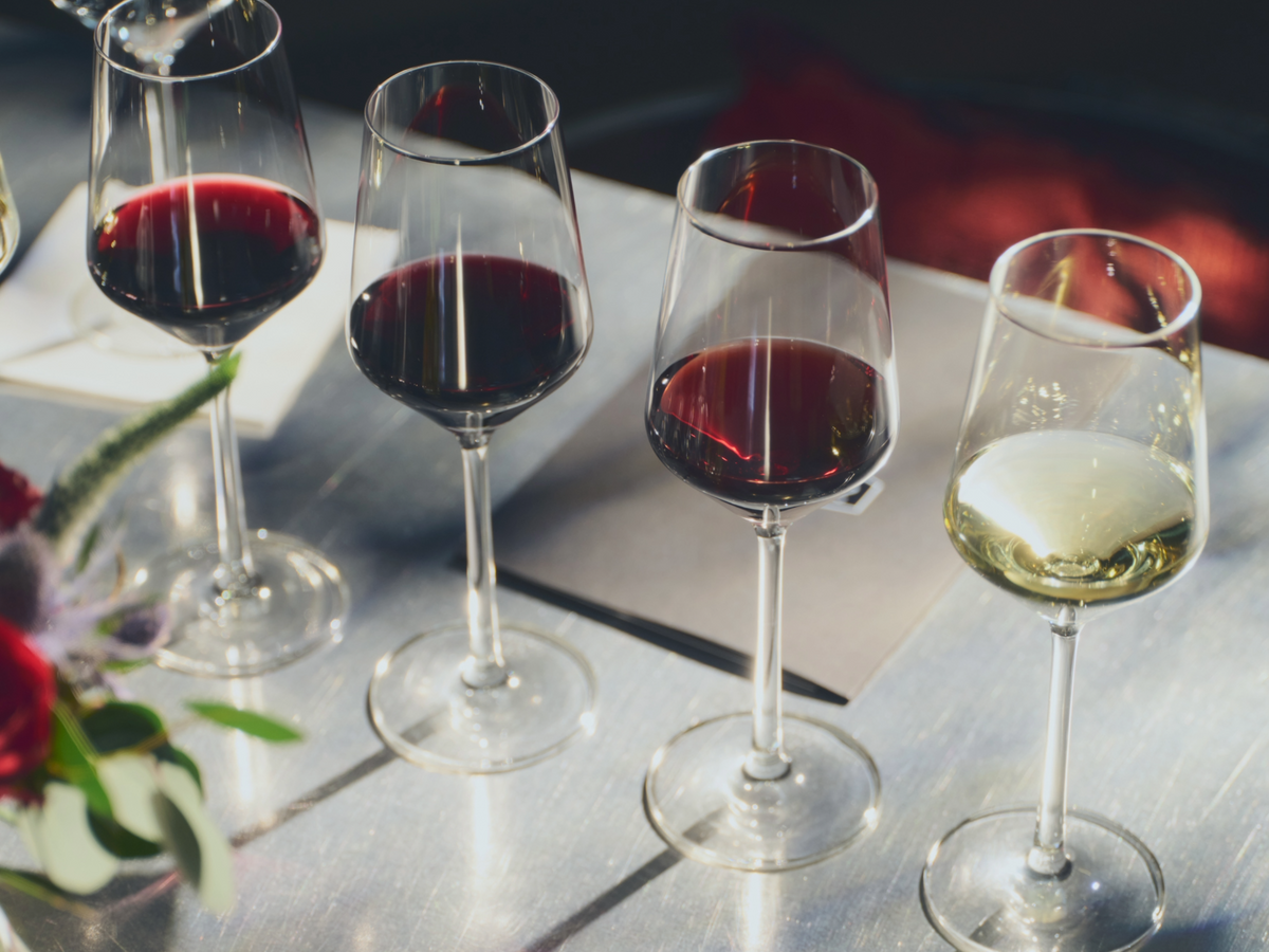 Four wine glasses on a metallic table, three filled with red wine and one with white wine, with a blurred background and flowers at the side.