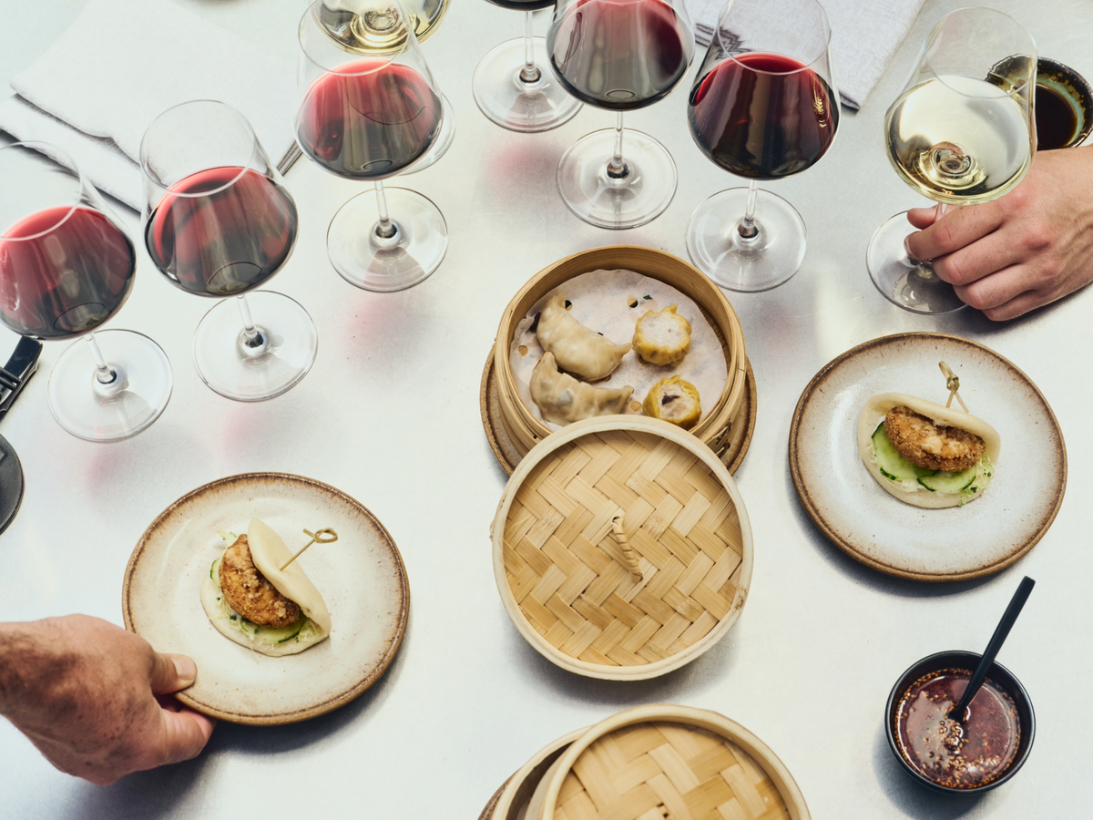 Overhead view of a dining table with plates of dim sum, bamboo steamers, wine glasses, and hands holding glasses and serving plates.