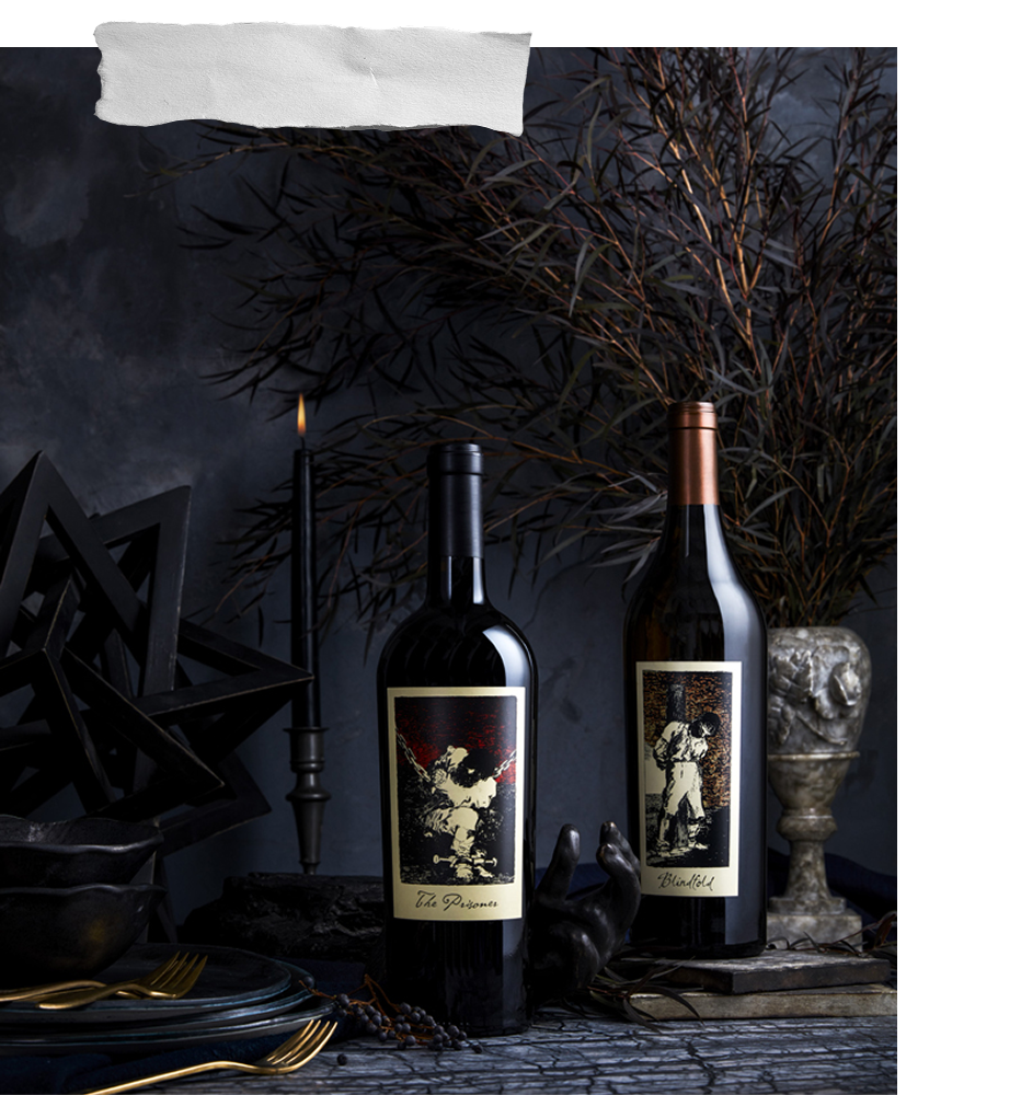 Two dark wine bottles with vintage-style illustrated labels are arranged on a dark tabletop with a candle, decorative objects, and dried branches in a moody, gothic-themed setting.